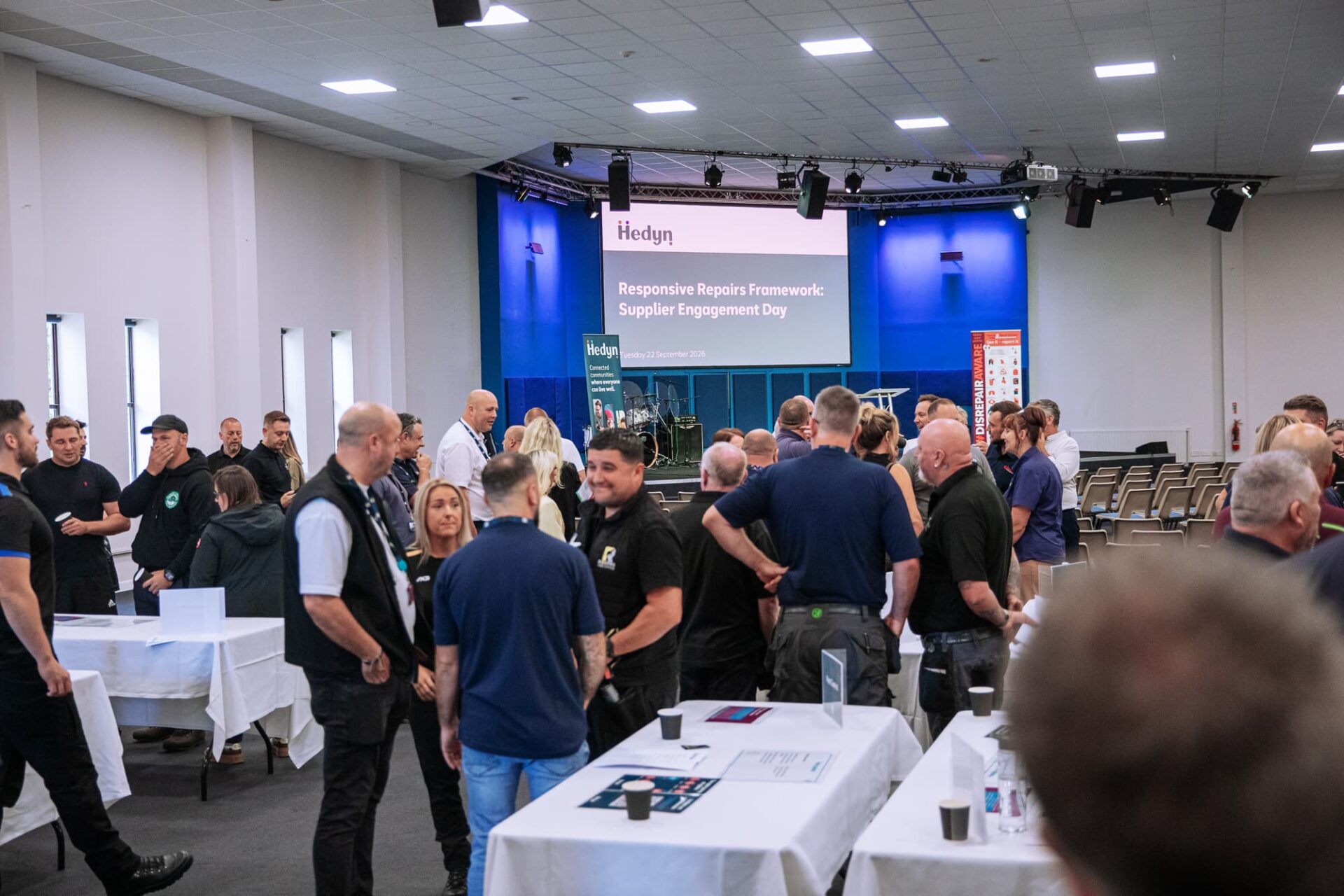 A large group of people at a conference, chatting, with a large presentation screen in the background.